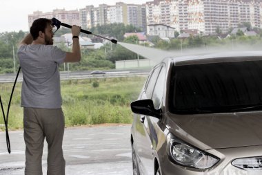 Gri renkli genç bir adam arabasını araba yıkamada yıkar. Kendi kendini temizleyen binek otomobiller için şehir servisi. Sakallı genç bir adam arabanın üzerine su döküyor, arabadaki kiri yıkıyor.