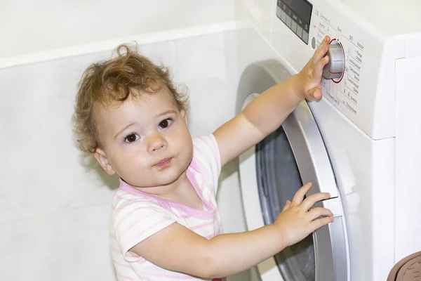 Lilla kaukasiska baby girl 1 år vrider på knappen på tvättmaskinen Stockfoto