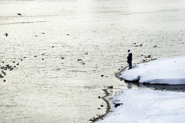 Couple man and woman stand on the snowy bank of the river and feed the ducks