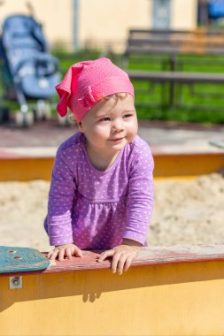 Mor elbiseli bir yaşındaki bir kız çocuğu neşeli bir şekilde kumluk bahçesine bakıyor. Bir çocuğun dikey portresi olan yürüyüş yapan mutlu bir beyaz çocuk.