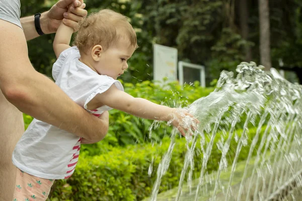 Menina brincando com água em uma fonte no parque. A criancinha molha as mãos na fonte. Papai caminha com uma criança no parque — Fotografia de Stock
