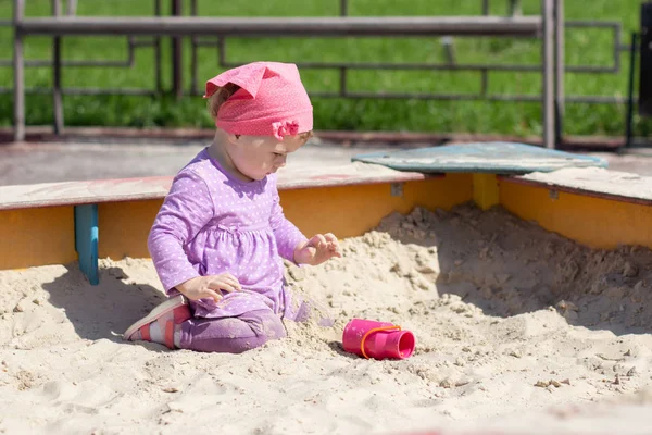 Το κοριτσάκι με το μωβ φόρεμα και το ροζ φουλάρι παίζει με μια κανάτα με παιχνίδια. Ευτυχισμένο Καυκάσιο παιδί 1 έτους για μια βόλτα, πορτρέτο ολόκληρου του παιδιού — Φωτογραφία Αρχείου
