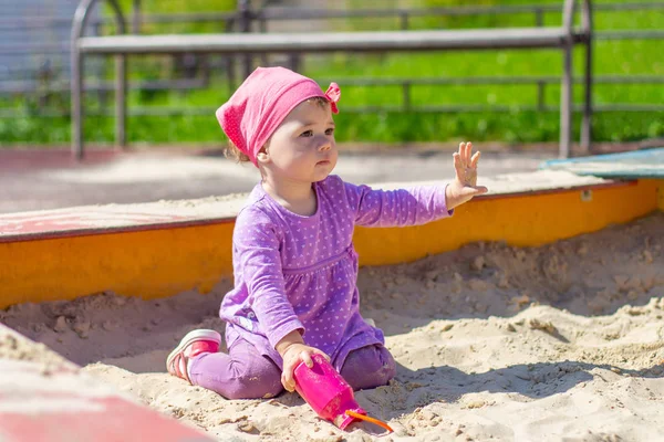 Baby girl in a purple dress plays with a toy pink jug outdoor. Happy Caucasian child 1 year old walking in the sandbox with raised hand