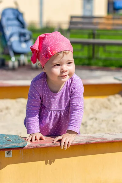 Mor elbiseli bir yaşındaki bir kız çocuğu neşeli bir şekilde kumluk bahçesine bakıyor. Bir çocuğun dikey portresi olan yürüyüş yapan mutlu bir beyaz çocuk.