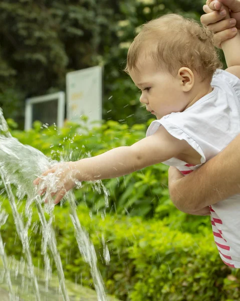 Bebê menina estende a mão para a fonte, o bebê brinca com jatos de água no parque. Uma criança em um passeio no parque brinca com água em uma fonte — Fotografia de Stock