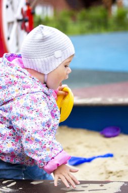 Çocuk kum havuzunda otururken çığlık atıyor. Şapkalı bebek ağzını açtı. Küçük kız bahçede oyun bahçesinde yürüyor.