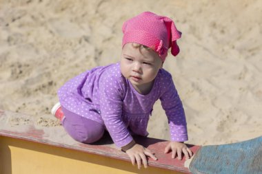 Leylak elbiseli ve pembe eşarplı küçük bir kız kum havuzunun kenarına tırmanıyor, güneşli bir gün.