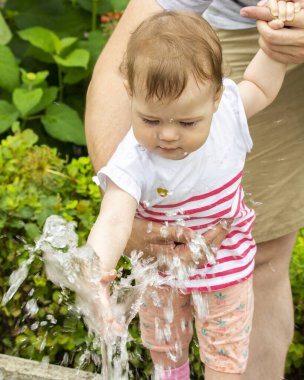 Küçük kız fıskiyede su püskürterek oynuyor. Parkta yürüyüş yapan bir çocuk suyla oynar, bebek çeşmeye tırmanır.