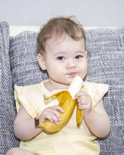 Klein meisje in het geel met een banaan in haar hand. Baby meisje reinigt banaan, baby eet exotisch fruit. Lastige blik. — Stockfoto