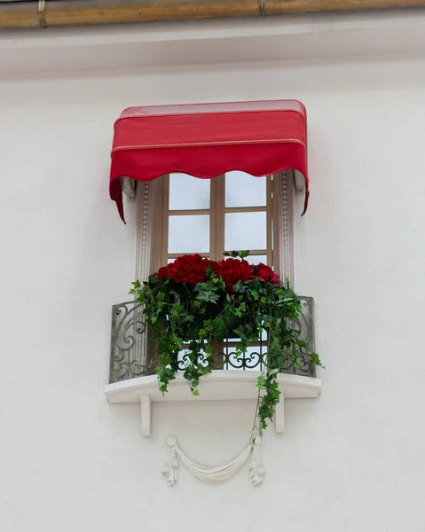Beautifully decorated window, French balcony with red flowers and a canopy. White wall of a house with a flower pot and molding