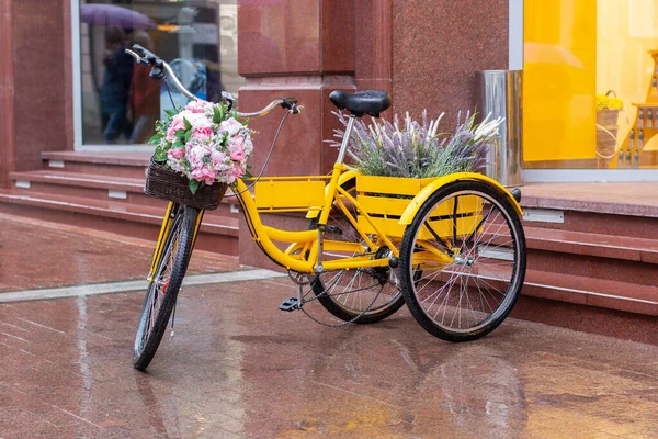 A romantic yellow bike stands on a wet marble pavement after rain. Bicycle with a basket of flowers, lavender and peonies. — Stock Photo, Image