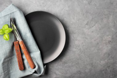 Masa ayarı. Gri beton arka plan üzerinde siyah plaka, çatal bıçak takımı ve peçete.
