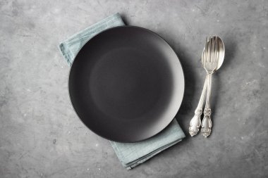 Öğle veya akşam yemeği konsepti. Masa ayarı. Siyah tabak, çatal bıçak ve peçete gri beton arka planda..