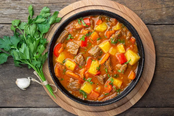 Goulash, beef stew or bogrash soup with meat, vegetables and spices in cast iron pan on wooden table. Hungarian cuisine. Rustic style. Top view.