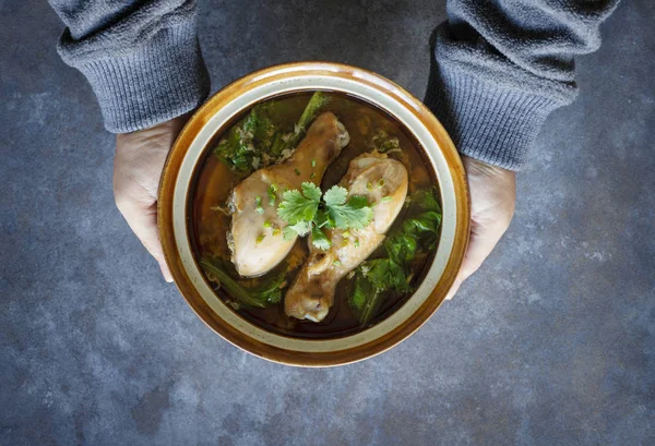 male hands holding portion of soup with chicken legs in brown ceramic bowl