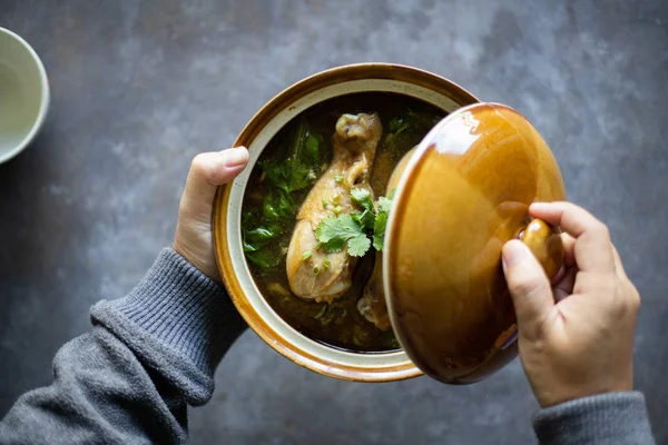 male hands holding portion of soup with chicken legs in brown ceramic bowl