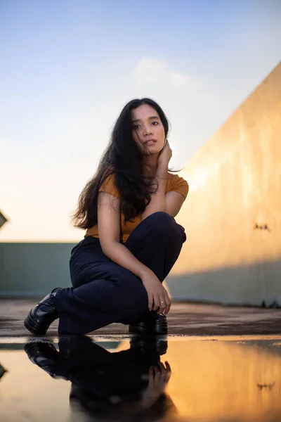 young woman in orange t shirt posing by puddle with reflection and evening sunlight