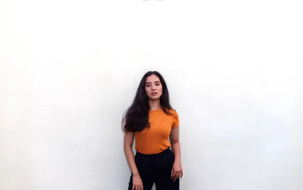 young woman in orange t shirt posing by wall