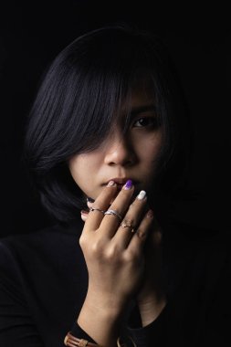 Asya kısa saçlı kız siyah uzun kollu gömlek ve metal ve deriden yapılmış takı giyiyor, siyah bir arka plan ve gölgeler üzerinde üzgün yüzler