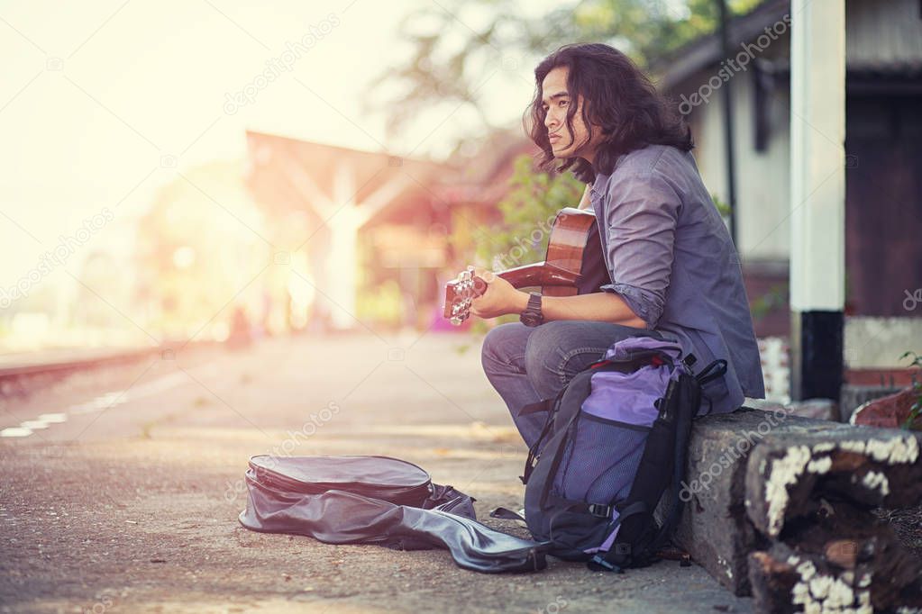Asian long haired man is playing the guitar while he is traveling. With a rustic background and trees Under the soft sunlight in the day
