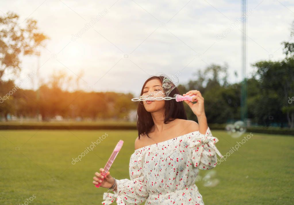 Attractive Asian young woman is enjoying blowing a soap bubbles in the public park with warm light 