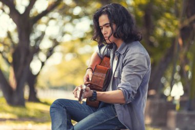 Asya uzun saçlı adam o seyahat ederken gitar çalıyor. Rustik bir arka plan ve ağaçlar ile gün yumuşak güneş ışığı altında