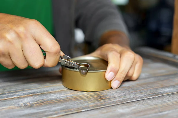 Mans hands open a tin can with an old rusty can opener with a metal handle. Canned food and a special knife. Selective focus.