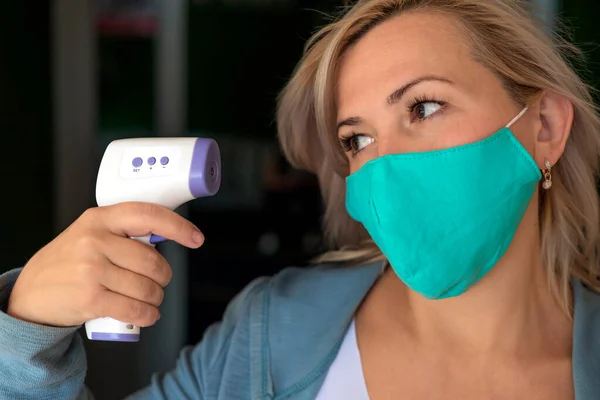 Tıbbi maskeli yetişkin bir beyaz kadın kendi ateşini kızılötesi termometreyle ölçüyor. Komik tıbbi konsept. - Stok İmaj