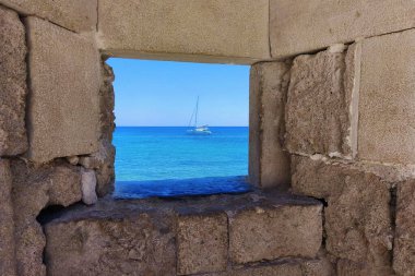 Antik duvardan deniz manzarası, seyahat ve tatil konsepti. Turkuaz deniz ve yelkenli yaz tatili. Okyanusa açılan taş pencere. Deniz. Rodos Adası Yunanistan - Eski kentteki kale.