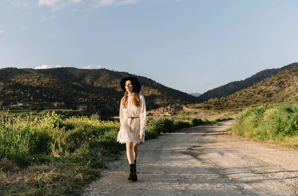 Young adorable girl in white dress and black hat, in spring, posing for camera. female portrait outdoor in mountain lake. Green fresh grass. Young unusual person enjoying nature