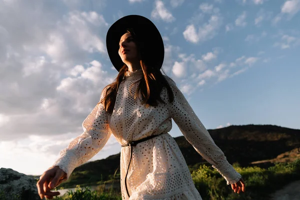 Young adorable girl in white dress and black hat, in spring, posing for camera. female portrait outdoor in mountain lake. Green fresh grass. Young unusual person enjoying nature