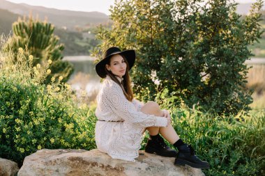 Beyaz elbiseli, siyah şapkalı, bahar aylarında kameraya poz veren sevimli bir kız. Dağ gölünde kadın portresi. Yeşil taze çimenler. Doğanın tadını çıkaran genç sıradışı kişi.