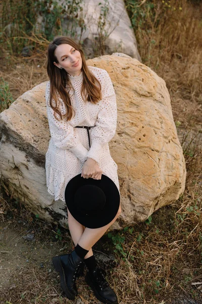 Young adorable girl in white dress and black hat, in spring, posing for camera. female portrait outdoor in mountain lake. Green fresh grass. Young unusual person enjoying nature