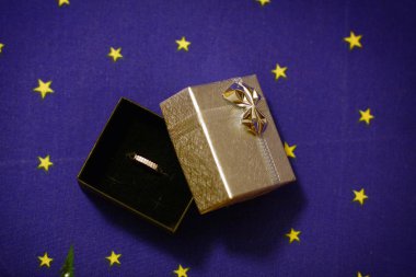 Hediye kutusunda elmas yüzük, mavi arka planda yay ve sarı yıldızlar.