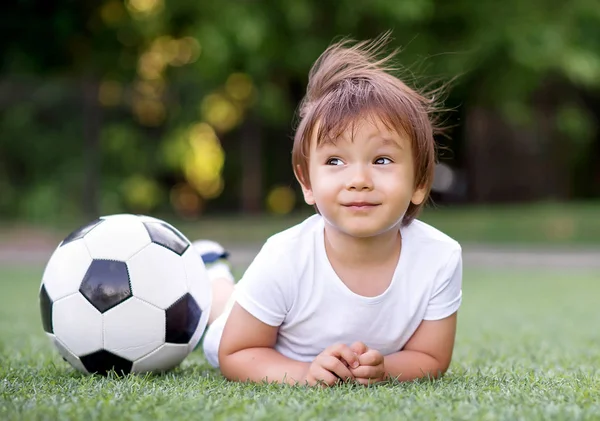 Criança jazendo na barriga no campo de futebol perto de bola de futebol e sonhando. O vento está a acenar com o cabelo do miúdo. Futuro conceito de desportista — Fotografia de Stock