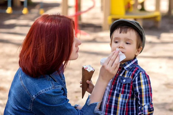 母は幼児の息子の顔を拭く。少年は屋外の散歩にワッフルコーンでアイスクリームを手に持っています。背景に子供の遊び場。マザーケアと夏の活動コンセプト — ストック写真