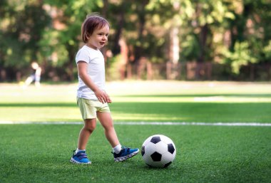 Oyun sırasında küçük futbolcu: spor üniforması küçük çocuk yaz günü açık havada futbol sahasında futbol oynuyor. Çocuk topa vurmaya hazır. Aktif çocukluk kavramı. Kopyalama alanı