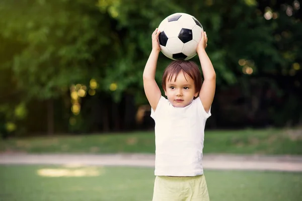 Pequeno jogador de futebol no campo de futebol ao ar livre: menino criança segurando bola acima da cabeça pronto para jogá-lo no dia ensolarado. Verão ao ar livre diversão e esportes para crianças conceito. Espaço para texto de cópia — Fotografia de Stock