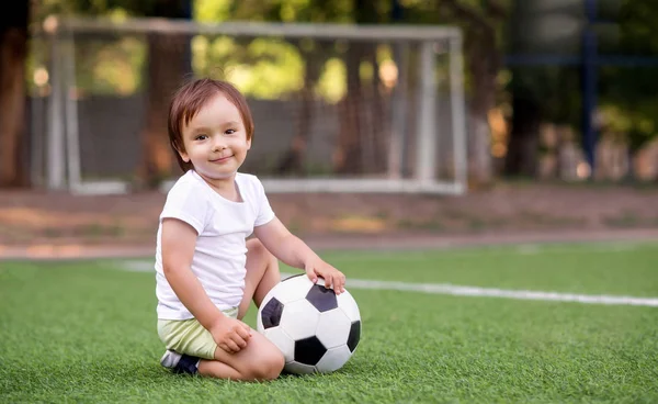 Pequeño niño en uniforme deportivo sentado con pelota de fútbol en el campo de fútbol al aire libre en el día de verano. Puestos de gol (redes de fútbol) en el fondo. Deportes infantiles y concepto de infancia activa. Espacio para texto de copia — Foto de Stock