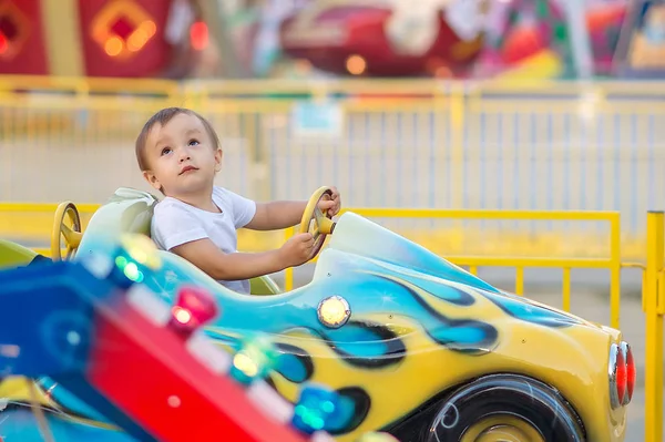 Ένα μικρό αγόρι που ιππεύει σε ένα αυτοκίνητο σε ένα χαρούμενο γύρο (καρουσέλ) στο θεματικό πάρκο και κοιτάζοντας πάνω. Φωτεινές απολαύσεις στο παρασκήνιο. Καλή ιδέα για την παιδική ηλικία — Φωτογραφία Αρχείου