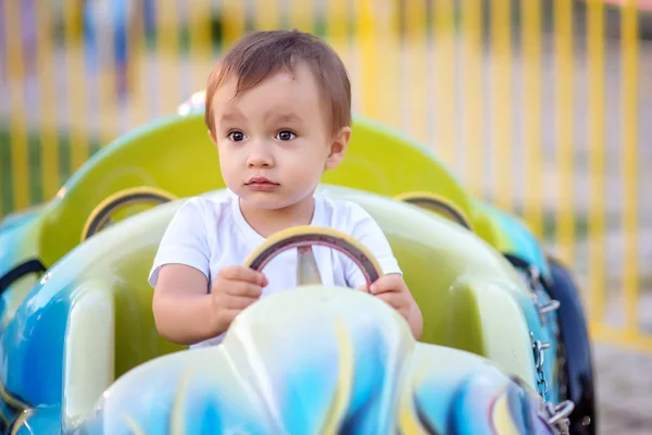 Pikkupoika istuu pienellä autolla karusellin päällä huvipuistossa. Poika pitelee rattia. Tulevaisuuden kilpa-ajaja käsite — kuvapankkivalokuva