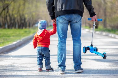 Babası ile yürüyüşe küçük yürümeye başlayan çocuk, arkasından görünüm. Babam mavi Kick Scooter tutarken çocuk, babasının el hodling. Bahar ve sonbahar açık havada aktivite kavramı