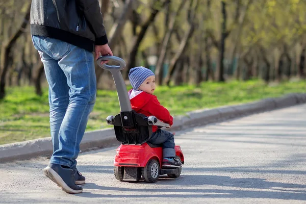 O pai empurra um carro vermelho com o filho criança montando-o ao longo de uma estrada de primavera. Árvores no fundo. Pai e filho em uma caminhada ao ar livre juntos. Espaço para texto de cópia — Fotografia de Stock