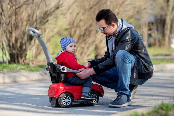 Father and little son talking outdoor in spring or autumn: kid is sitting on a red push car while father has squatted down in front of car and explains to him road safety rules. Traffic law for children concept — ストック写真