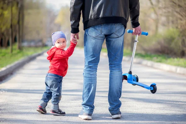 Baba ve yürümeye başlayan oğul bir yürüyüşe el tutarak, arkadan görünüm için gidiyor. Babam küçük mavi tekme scooter taşıyor, oğlum yarısını geri döndü ve geri bakıyor. Aile eğlence açık havada kavramı
