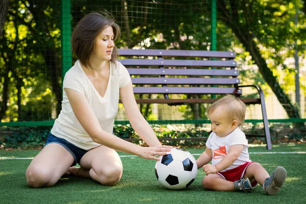 若いママと赤ちゃんはサッカー場に座ってお互いにサッカーボールをロール。母と息子は一緒に遊んでいます。夏の家族スポーツ活動コンセプト — ストック写真