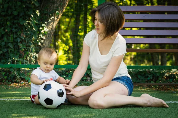Ein kleiner Junge schaut sich den Fußball an und erkundet ihn, während er neben seiner Mutter auf dem Fußballplatz sitzt. Kleinkind Sohn spielt mit Mama im Freien. Aktives Familien- und Kinder- und Sportkonzept. — Stockfoto