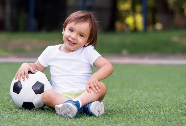 Criança sorrindo menino sentado com pernas cruzadas no campo de futebol no dia de verão com bola de futebol. Criança feliz jogando futebol ao ar livre. Conceito de infância ativa — Fotografia de Stock