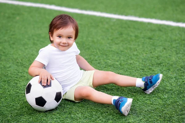 Pequeno jogador de futebol sentado em um campo de futebol verde com uma bola e sorrindo. Retrato de uma criança feliz brincando com uma bola ao ar livre. Conceito ativo de esportes de infância e verão — Fotografia de Stock