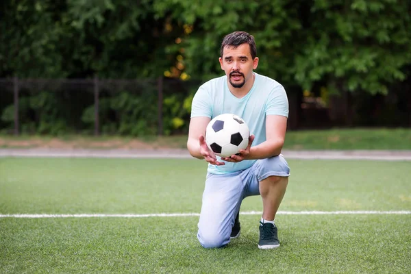 片膝に立ち、サッカーボールをカメラに向かって話すサッカーコーチや体育教師の肖像画。緑のサッカー場でボールでひざまずく中年の男 — ストック写真
